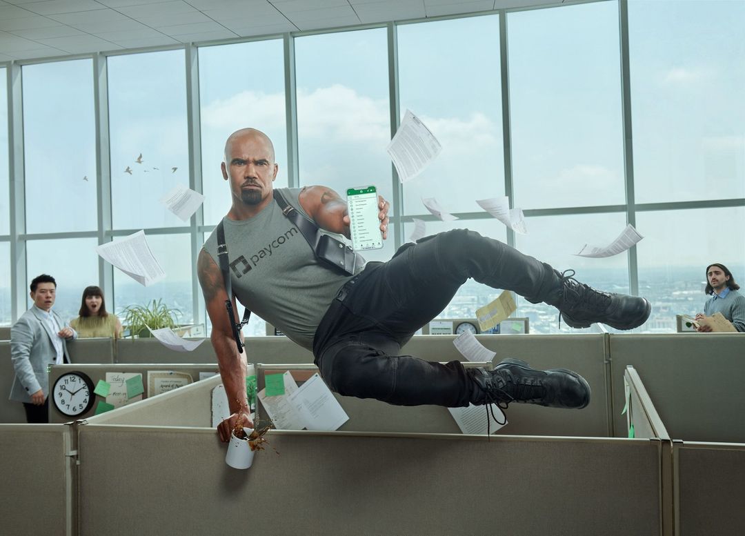 An adult male leaping over a cubicle in a modern office, holding a smartphone and wearing tactical gear, with surprised coworkers in background.